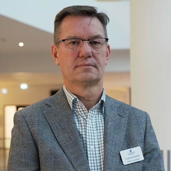En person iklädd en grå kavaj med en namnbricka står inomhus i en modern byggnad med vit interiör och diskreta belysningsarmaturer i bakgrunden.