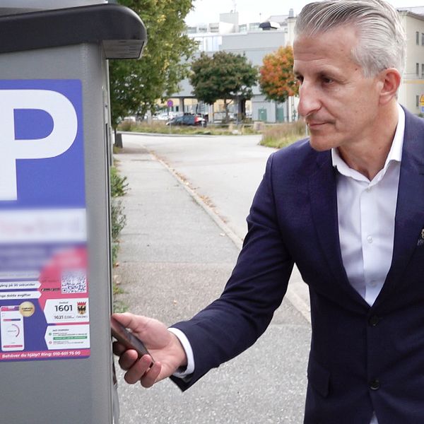 Kemal Hoso (S), ordförande i exploateringsnämnden i Örebro står nära en parkeringsautomat med en P-skylt på framsidan. Han håller en nyckel eller kort i handen och ser ut att interagera med automaten. I bakgrunden syns en grönskande gata med byggnader och träd.