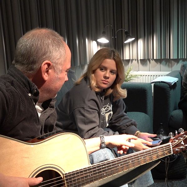 Tre musiker sitter i två soffor. Man spelar gitarr.