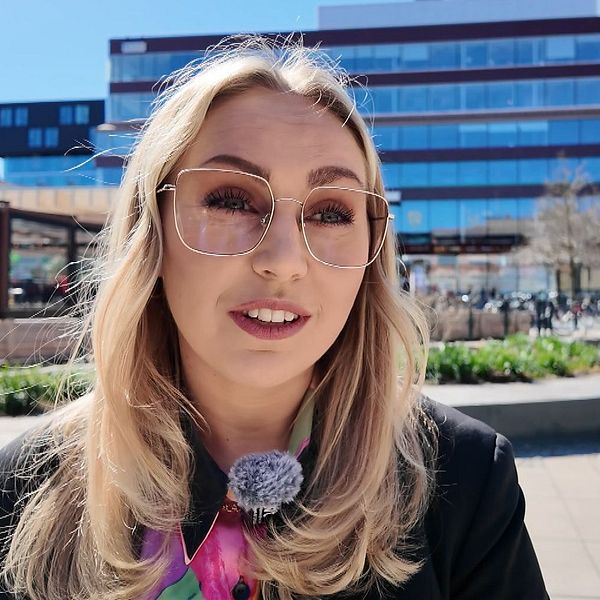 Kvinna med långt blont hår står utomhus i en stadsmiljö med moderna byggnader i bakgrunden.