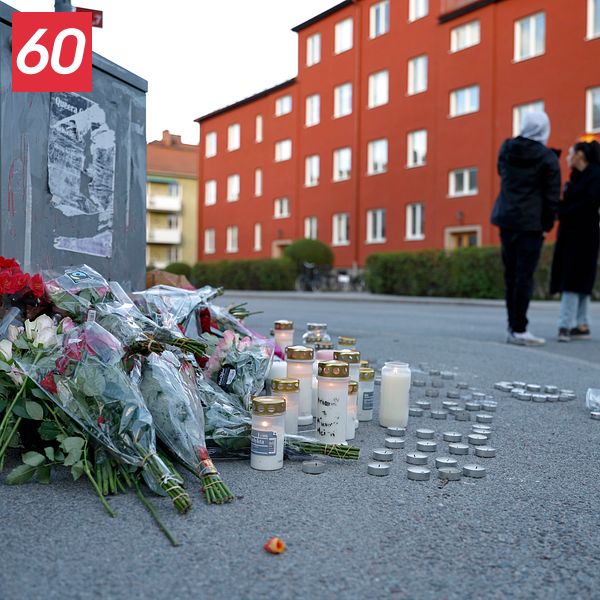 Blommor och tända ljus på trottoar, tre personer samlade i bakgrunden vid rött bostadshus.