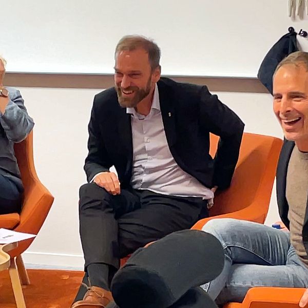 Tre personer sitter på orange fåtöljer under en diskussion eller paneldebatt. Det finns en liten kaffebord med en skål snacks och två flaskor med vatten i förgrunden.