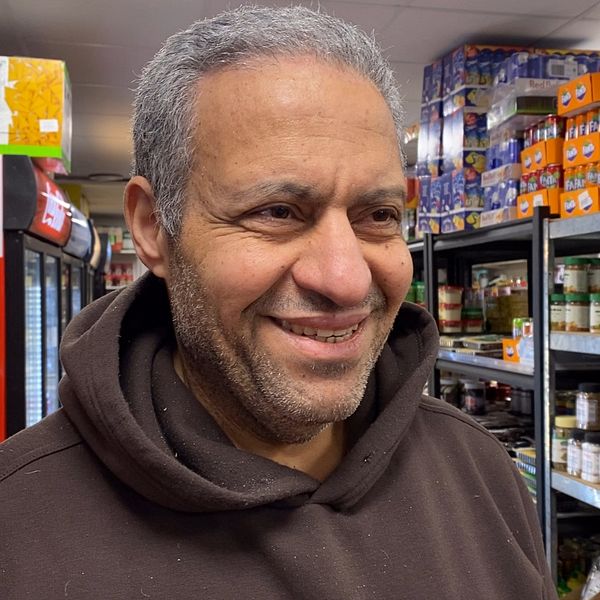 Ameen Talal står i sin butik i Lagersberg, som . Bakom honom syns hyllor med konserver och läskburkar. Han ser glad ut.