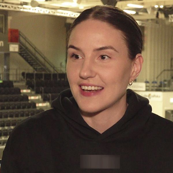 Ung kvinna i svart tröja står i en tom idrottsarena, läktare och basketplan syns i bakgrunden.
