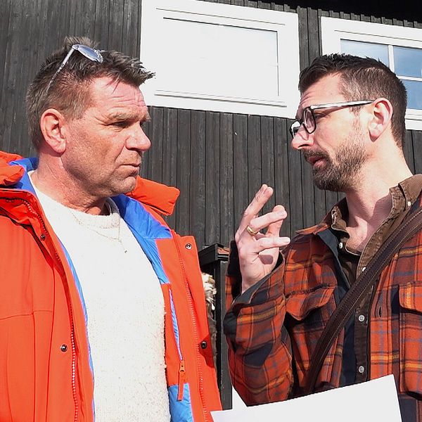 Två personer diskuterar utomhus framför ett mörkt trähus i solen.