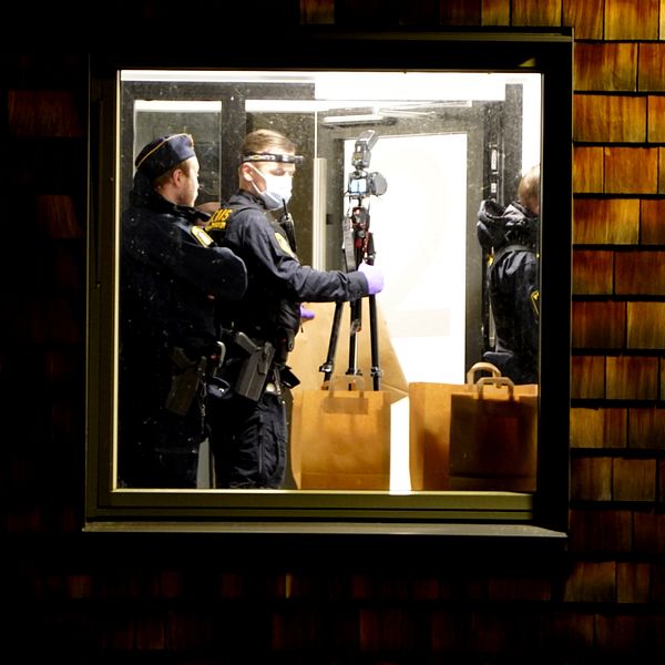 Poliser iklädda skyddsutrustning står inne i ett rum som syns genom ett fönster. De håller i en kamera på ett stativ och verkar förbereda sig för att dokumentera en scen, med en brun väska placerad på golvet. Bakgrunden består av en mörk vägg med trätak.