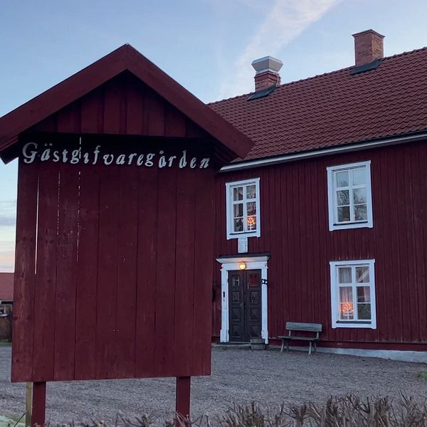 En röd timrad byggnad med vita fönster, tydligt markerad som ”Gästgivargården” på en skylt framför huset. Byggnaden har ett traditionellt utseende med två skorstenar och ligger i en lantlig miljö.