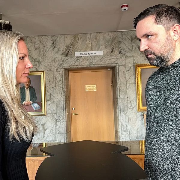 Två personer står vid ett bord, vända mot varandra. Rummet har en marmorvägg och innehåller två målningar på väggarna. En dörr är synlig i bakgrunden, och det finns en skylt ovanför.
