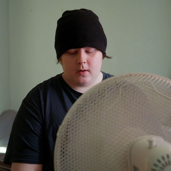 Gustaf sitter på säng med svart tröja och mössa, en fläkt syns i förgrunden.