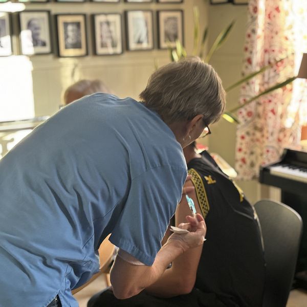 En person får en vaccination i en inomhusmiljö med en lugn atmosfär. Den som administrerar vaccinet står bakom och använder en spruta, medan den vaccinerade personen sitter ner och har på sig en svart t-shirt. I bakgrunden syns en piano samt fotografer på väggarna.