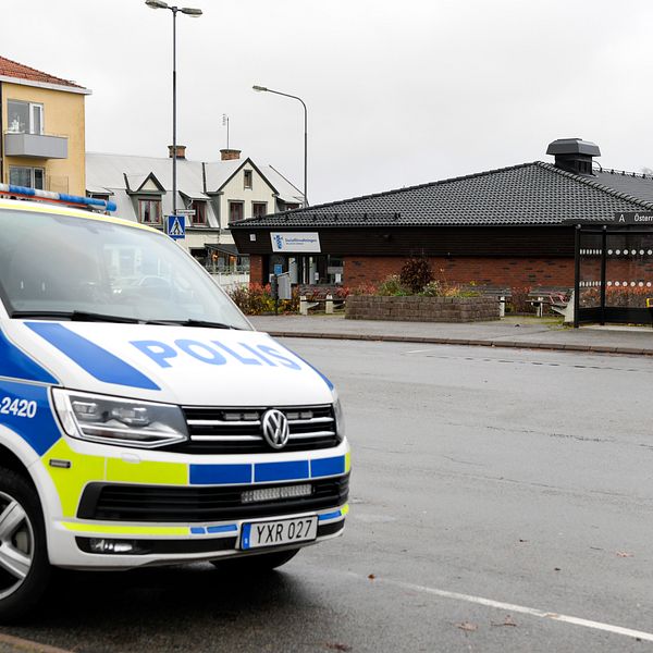 En polisbil står parkerad vid en busshållplats i en stadsområde, med en byggnad i bakgrunden, under ett molnigt väder.