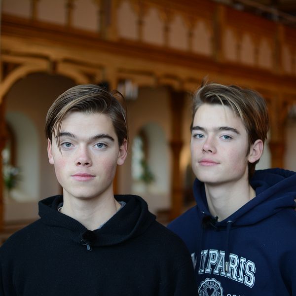 Två personer står i en byggnad med inredning som tyder på en historisk eller religiös miljö, med trädetaljer och valv i bakgrunden.