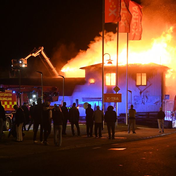 En nattlig scen där flera personer observerar en byggnad som brinner intensivt. Röd och orange eldslåga syns på taket och en brandbil är parkerad i närheten. Brandmän arbetar för att släcka elden.