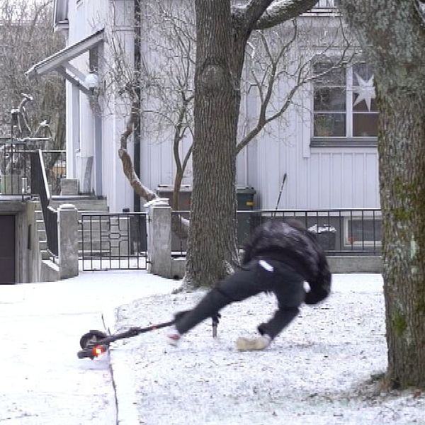 En person i svart kläder faller framåt på en snötäckt gård, nära en stor trädbas, med en bostad i bakgrunden och en basketkorg. Området ser kallt och vintrigt ut.