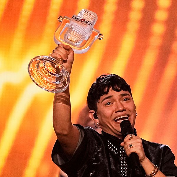 Person håller upp en Eurovision-trofé framför en lysande orange bakgrund.