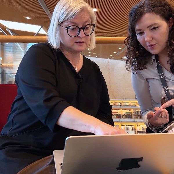 Två personer sitter vid ett bord i en biblioteksmiljö och tittar på en bärbar dator. Den ena personen pekar på skärmen medan den andra skriver. I bakgrunden syns en öppen yta med stolar och ett biblioteksliknande rum.