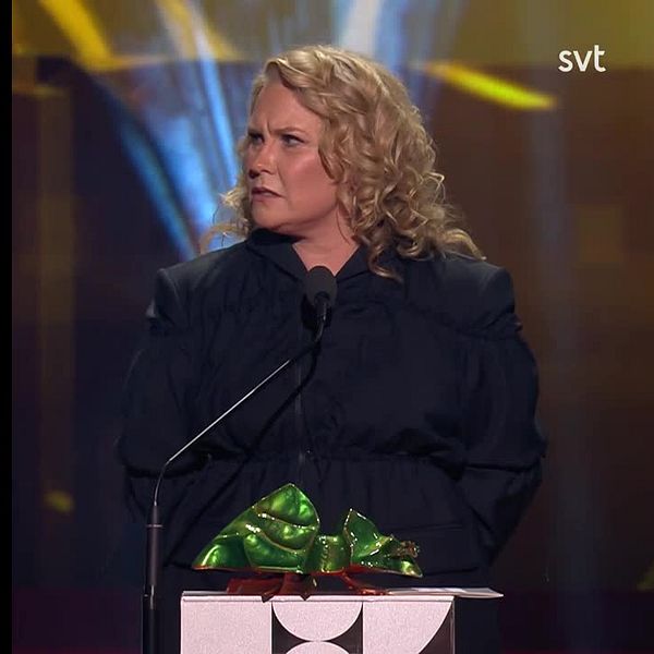 En person på scenen som står vid en mikrofon och håller i en festlig present insvept i grönt och guld. Bakgrunden har en färgglad belysning som skapar en festlig atmosfär.