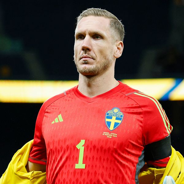 Svensk landslagsmålvakt i röd tröja tar av sig en gul jacka på fotbollsarena.
