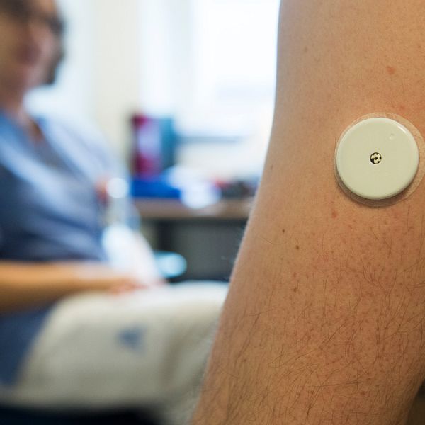 En arm med en medicinsk sensor fäst på huden, i bakgrunden en person i vårdmiljö.