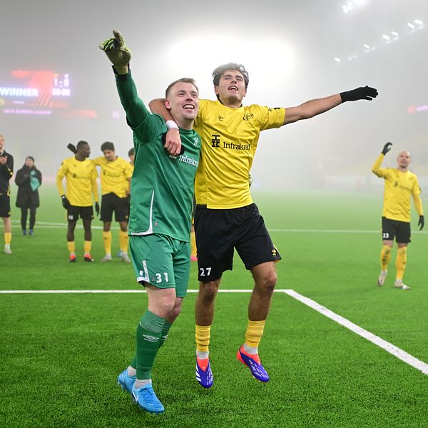 Två Elfsborgsspelare firar en seger på fotbollsplanen, omgivna av dimma. Den ena spelaren, i grön målvaktsdräkt, håller i den andra spelaren, som bär en gul tröja. I bakgrunden syns lagkamrater som deltar i firandet.