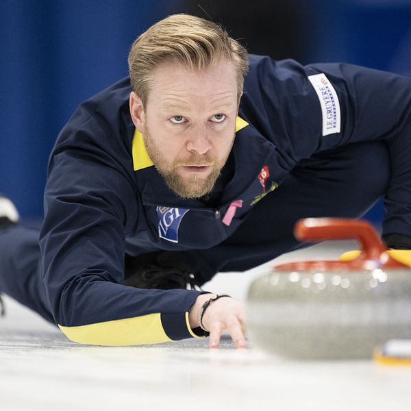 En curlingspelare ligger på knä på isen, sträcker sig fram mot en curlingsten med en snabb rörelse. Denna spelare bär en mörk jacka med gula detaljer och visar en koncentrerad hållning, med fokus på spelet. Curlingstenen ligger nära spelaren, på väg mot målet. Bakgrunden är suddig, vilket framhäver spelaren och stenen.