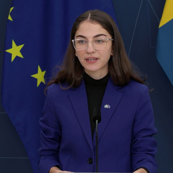En person står vid ett podium under en presskonferens, klädd i en blå kavaj. Bakgrunden består av svenska och europeiska flaggor.
