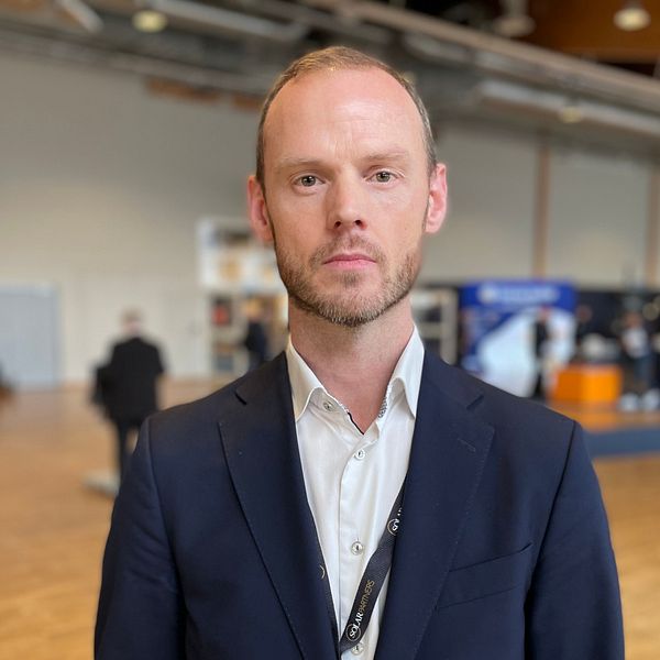 En person i kostym står i en konferenslokal med andra deltagare i bakgrunden.