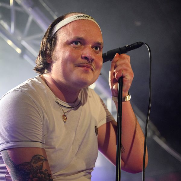 En manlig sångare i en vit t-shirt står vid en mikrofon med en scenbelysning som skapar en dramatisk atmosfär. Han har på sig en armbandsklocka och en halskedja, och hans arm visar tatueringar, vilket bidrar till den musikaliska stämningen.