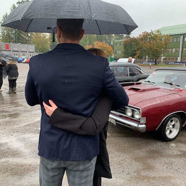 En man i en mörk kavaj håller om en kvinna under ett paraply, medan de står vid en röd veteranbil. Det regnar och flera personer syns i bakgrunden, som också är klädda för vädret. Området är en parkeringsplats omgiven av träd och byggnader.