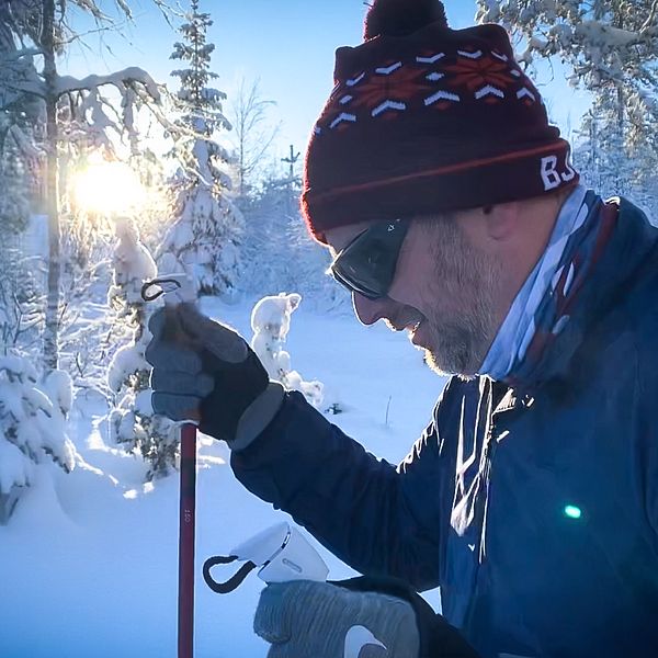 En man med mössa och svarta heltäckande glasögon åker skidor i en snötäckt skog