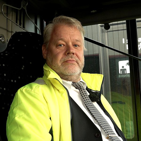 En person i en gula reflexjacka sitter vid ratten i en buss. Bakom syns bussens interiör och ett betalningssystem med kortläsare.