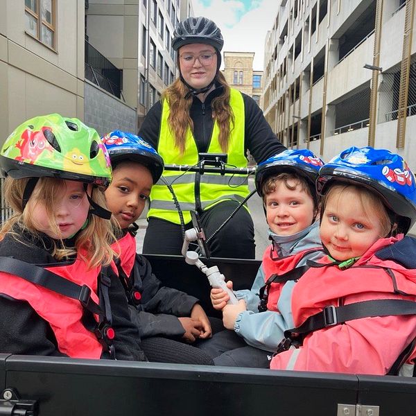 Flera barn med cykelhjälmar färdas i en lådcykel, vuxen med reflexväst cyklar bakom.