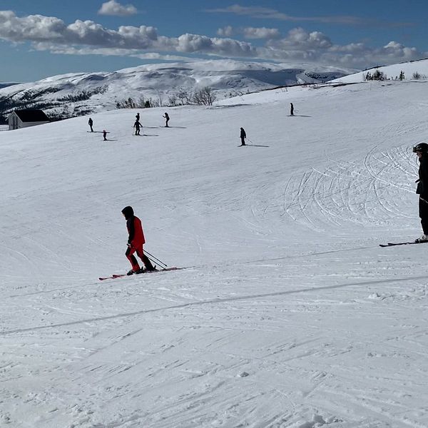 Skidåkare på en snöklädd backe i fjällen under en klarblå himmel, med flera personer utspridda i bakgrunden som åker skidor.