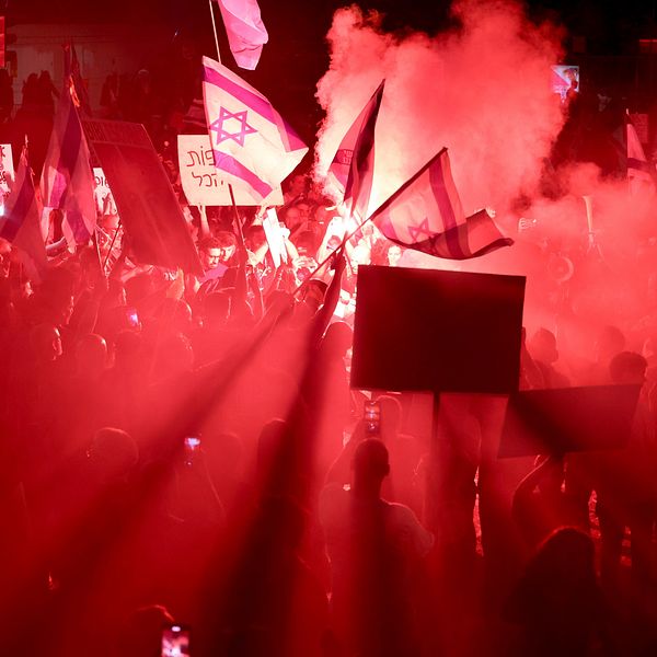 Demonstranter i Tel Aviv upplyst i rött ljus och rök viftar med israeliska flaggor och skyltar.