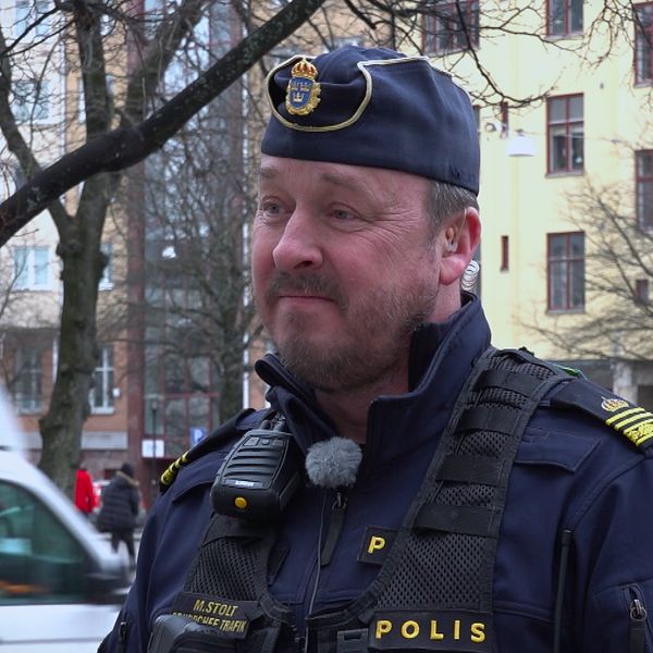 En polis står i en stad, med byggnader och ett vitt fordon i bakgrunden.