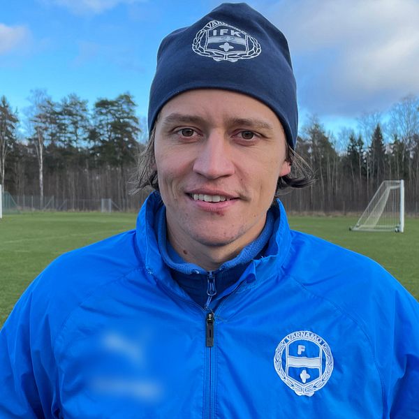 En person i en blå sportsjacka och mössa står på en fotbollsplan, omgiven av träd och två fotbollsmål i bakgrunden. Det är en klar dag.