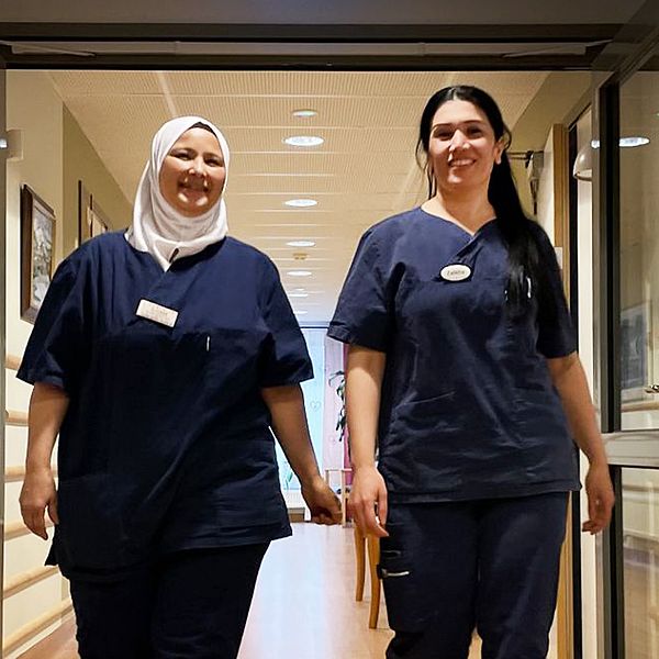 Kvinnorna Ilham och Zalikha i blå arbetskläder går i en korridor på ett äldreboende i Säffle