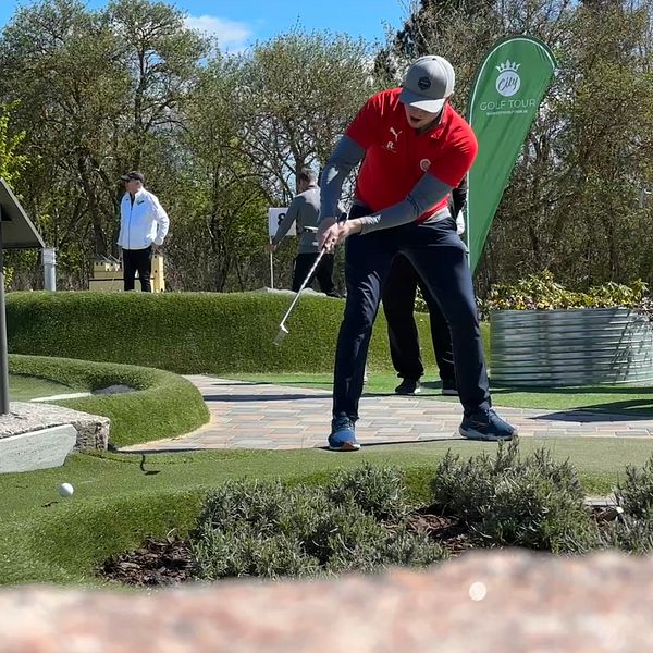 Person i röd tröja slår ett slag på en utomhusbana för minigolf, andra personer i bakgrunden.