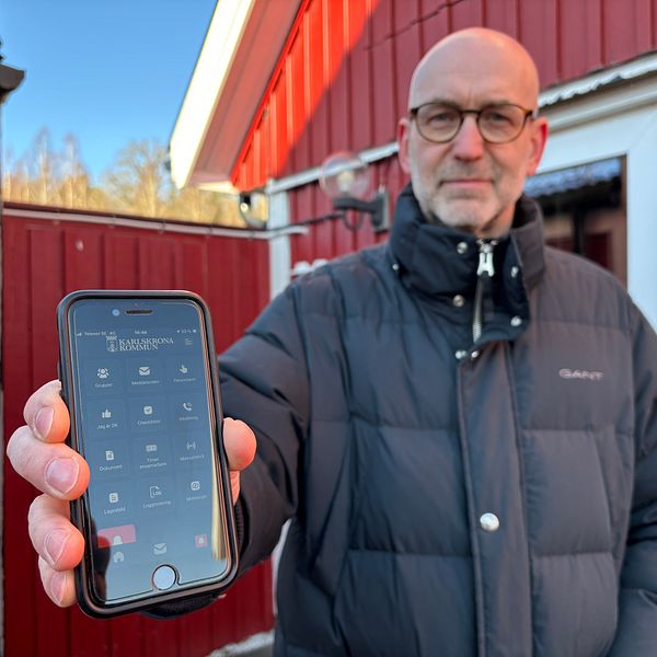 Person, Anders Viklander, i svart jacka visar mobiltelefon som har en krisinformationsapp installerad mot kameran utanför rött hus.