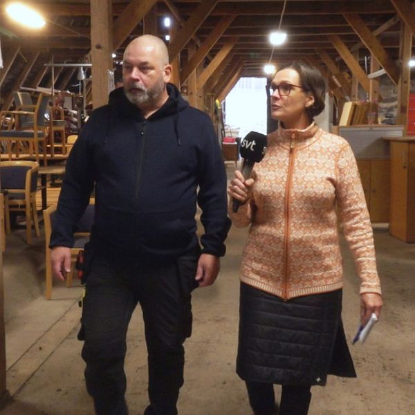 En man och en kvinna går tillsammans genom ett stort förråd, omgivna av stolar och bord. Kvinnan håller en mikrofon och talar. En stor, öppen lokal med träbjälkar i taket.
