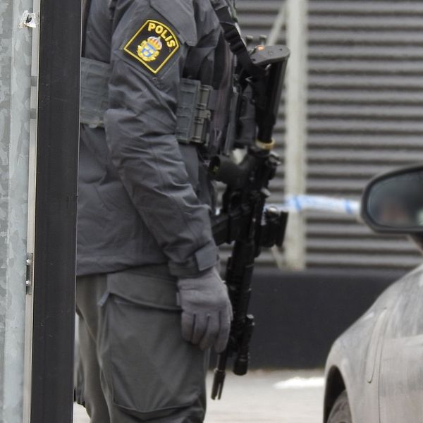 Bild på en polis i uniform med automatvapen. Han syns från halsen och knäna. Bredvid honom står en svart bil.