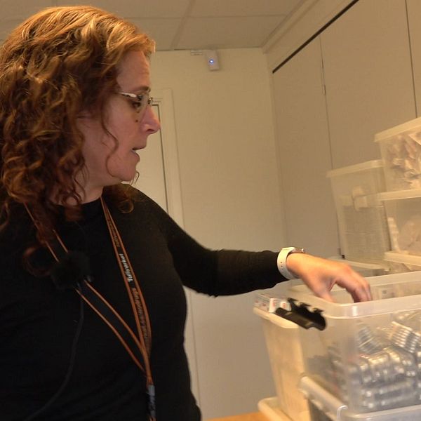 En person står vid ett bord i ett rum fyllt med förvaringslådor, och räcker ner en hand mot en öppen plastbehållare med tablettkartor.