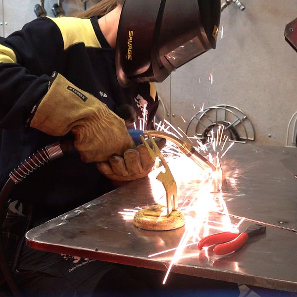 En person svetsar på ett metallstycke, vilket skapar gnistor. Personen bär skyddshandskar och en svetsmask, och arbetar på en metallplatta som är placerad på ett arbetsbord. I bakgrunden syns verktyg samt svetsutrustning.