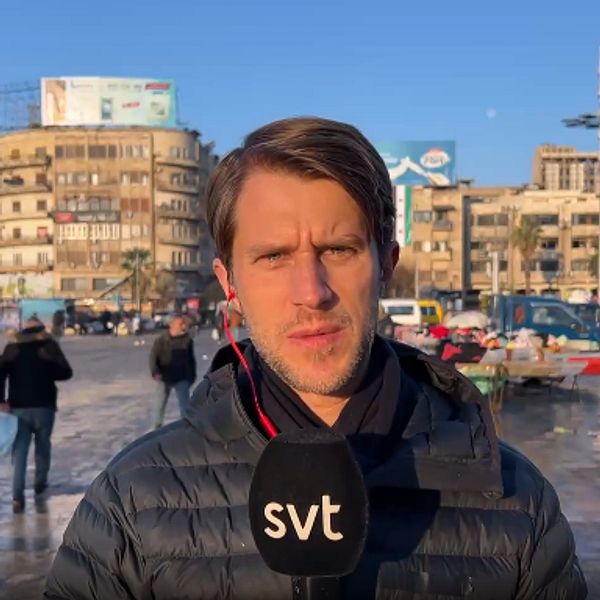 Thomas Thoren står på ett torg och rapporterar med byggnader i bakgrunden
