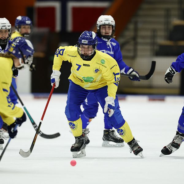 Svenska och norska bandyspelare tävlar på isen med Norgeflaggan i bakgrunden.