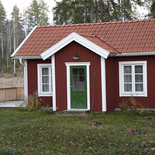 Röd stuga med vita knutar och gröna dörrar, omgiven av skog och gräsmatta.