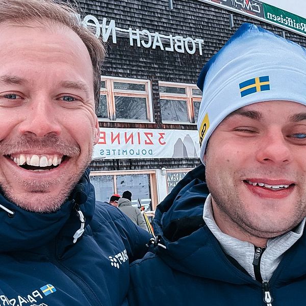 Två personer i vinterkläder står utomhus framför en byggnad med skyltar på ett främmande språk. Det är snö på marken och taket, vilket tyder på kallt väder.