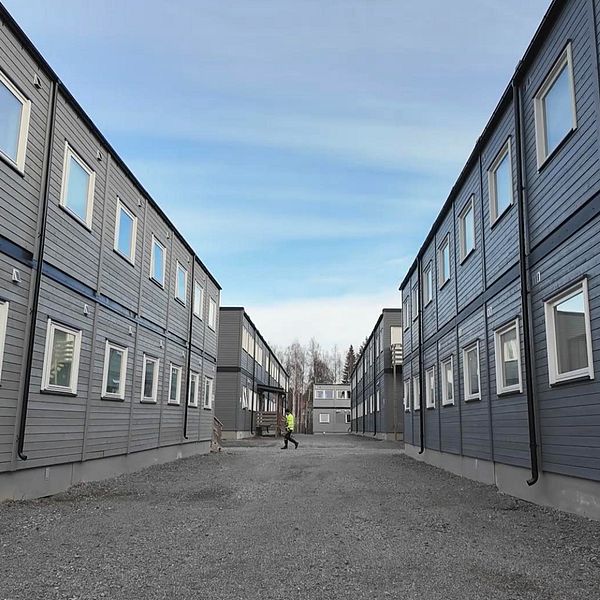 Synlig väg mellan två rader av gråbyggda modulhus med fönster, under en ljusblå himmel. En person går längs vägen mellan husen.