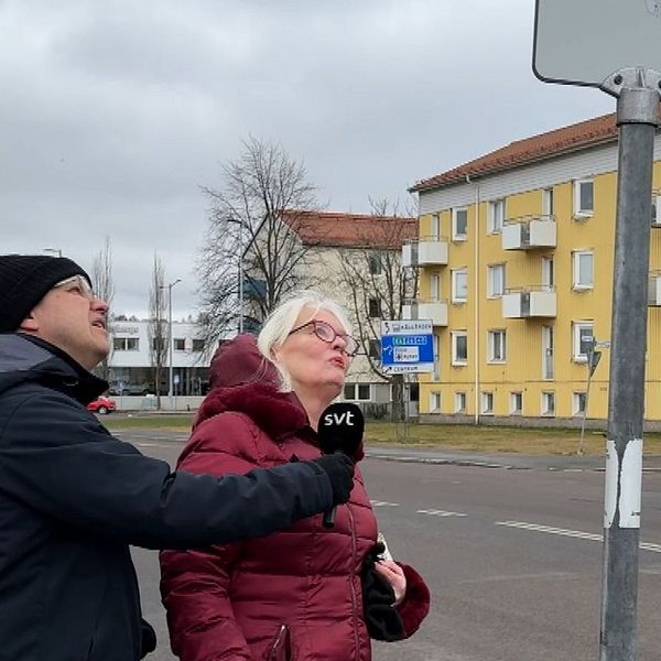 En man och kvinna tittar på en busskylt vid en gata med ett gult bostadshus i bakgrunden.