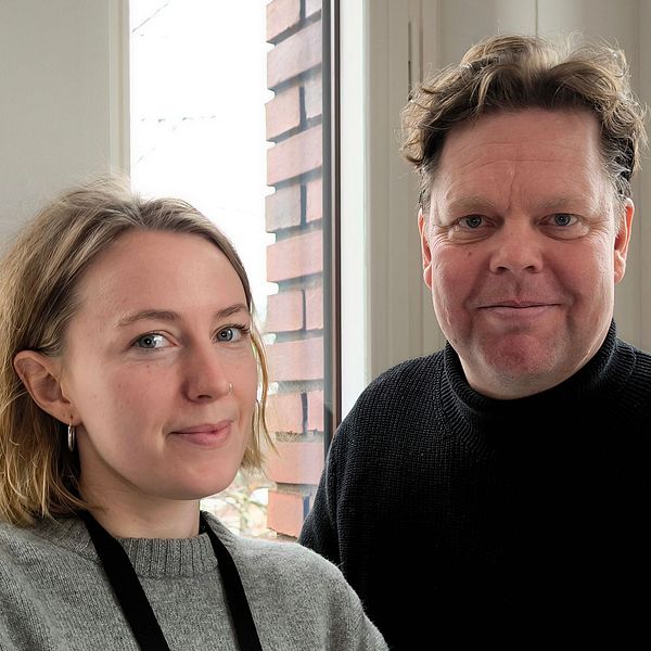En kvinna med kort, ljust hår och en grå tröja står bredvid en man med mörkt hår som bär en svart tröja. De står vid ett fönster med ljusinsläpp och en bakgrund av tegelvägg.